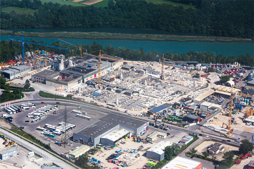 Aerial view of a large industrial yard with tower cranes, warehouses, stacked materials and rows of parked buses, beside a river with wooded hills behind.