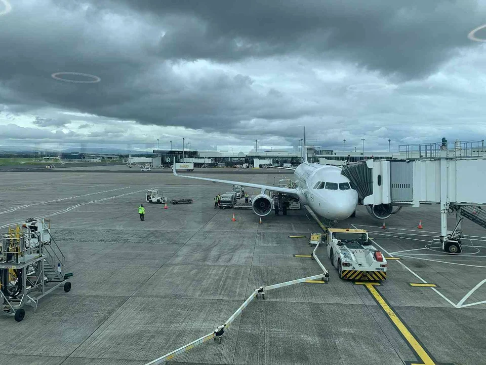 Passenger jet parked at a boarding bridge on a wet airport apron under heavy grey clouds, with a pushback tug and ground crew nearby.