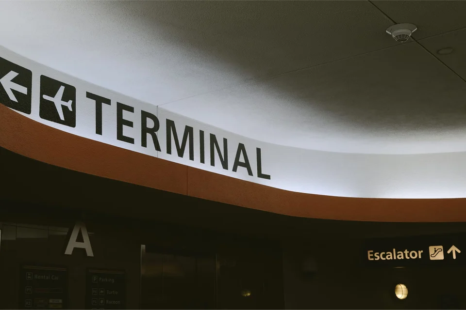 Illuminated airport sign reading 'Terminal' with a left arrow and an aeroplane symbol on a curved overhead wall, with an 'Escalator' sign below on the right.