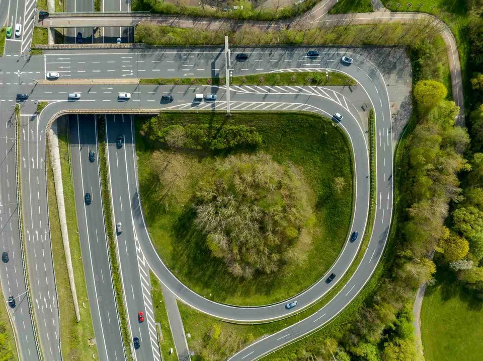 Aerial view of a highway interchange, where a looping ramp curves around a grassy patch of trees between multi-lane carriageways.