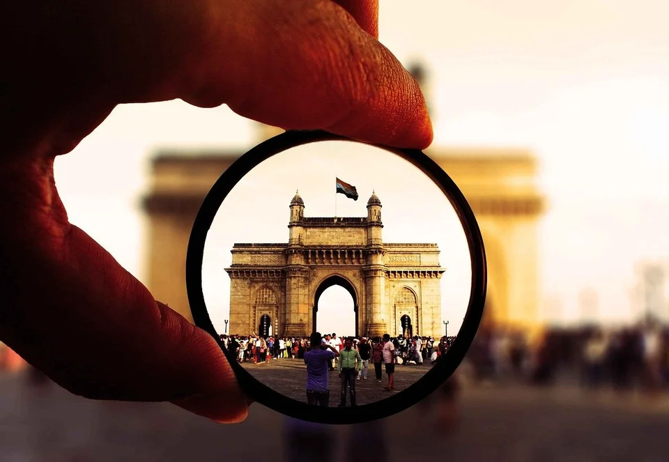 A hand holding up a round lens that brings the Gateway of India into sharp focus, with crowds of visitors in front of the arch and the scene blurred around it.