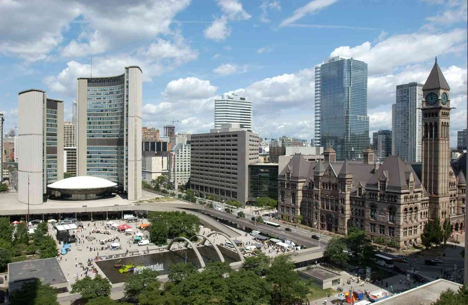 City skyline on a sunny day with two curved high-rise towers around a domed building, a busy public square with a pool, and an older stone building with a clock tower.