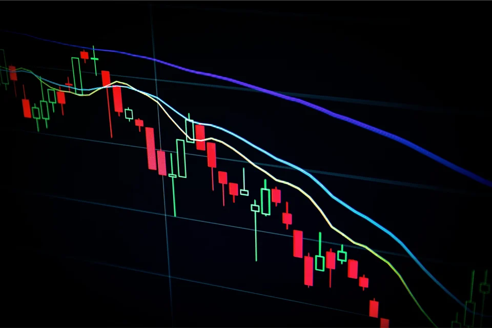 A candlestick price chart on a dark screen, with red and green candles and coloured moving-average lines sloping downward.
