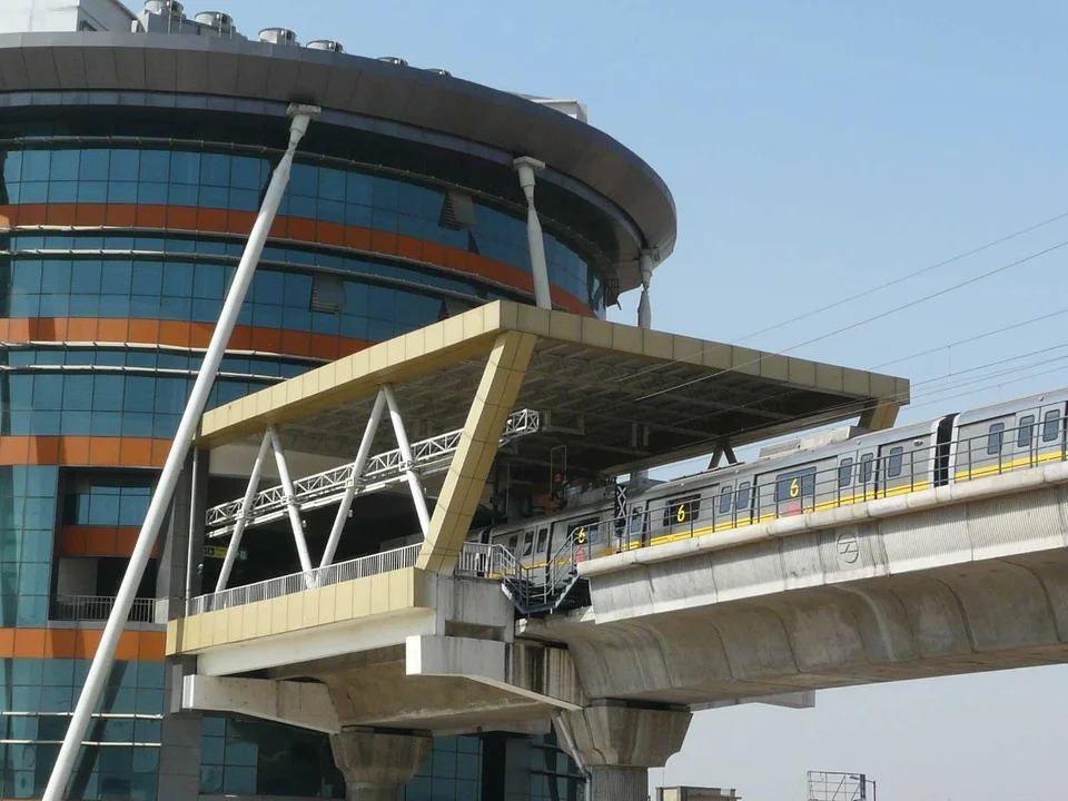 A metro train on an elevated track beside a station walkway and a curved, glass-fronted building with orange bands.