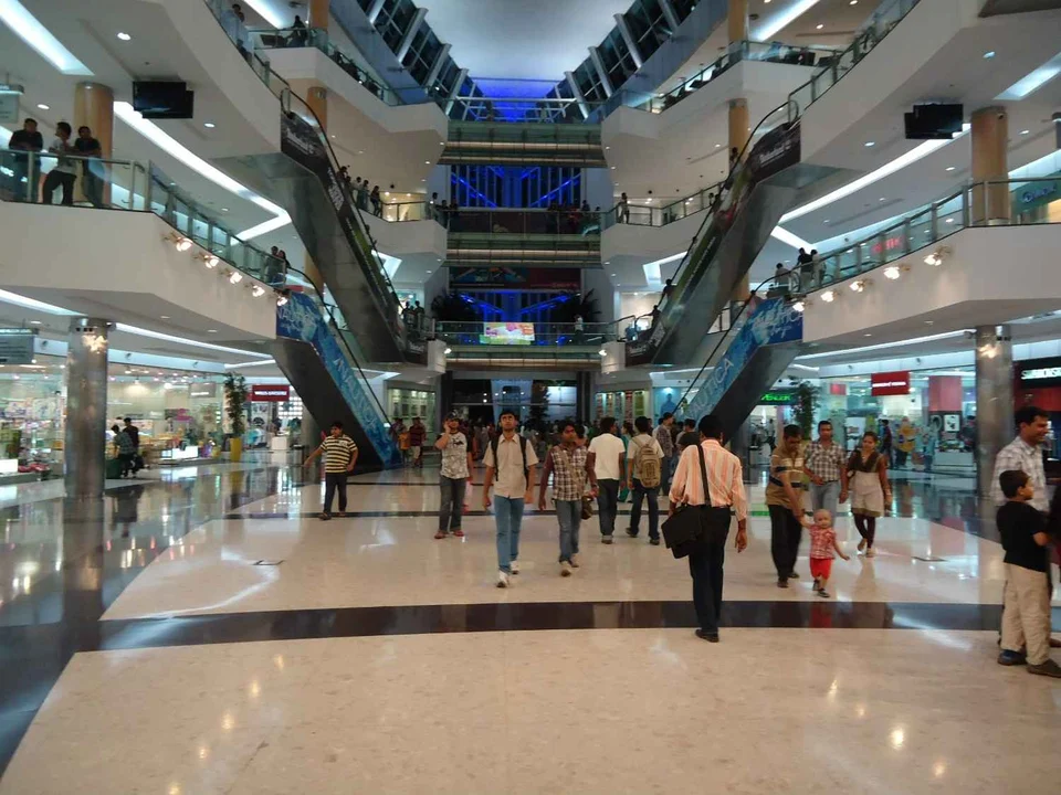Shoppers walking across the polished floor of a busy multi-storey shopping mall, with escalators rising through a central atrium.
