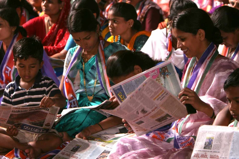 A group of women in saris and young children sitting together on the floor, reading newspapers.
