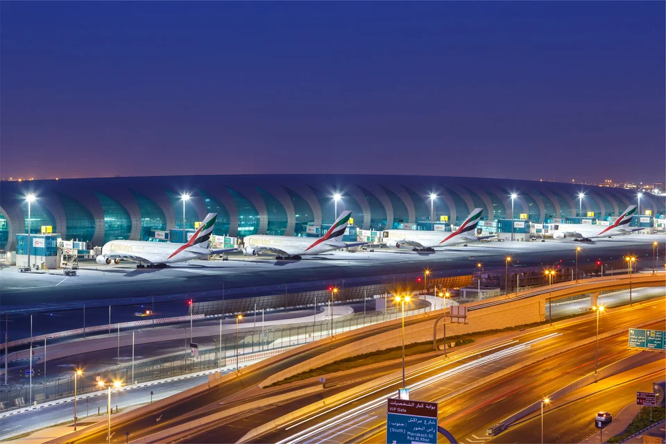 Large airport terminal at dusk with a row of wide-body jets parked at its gates, and a lit multi-lane road with traffic light trails in the foreground.