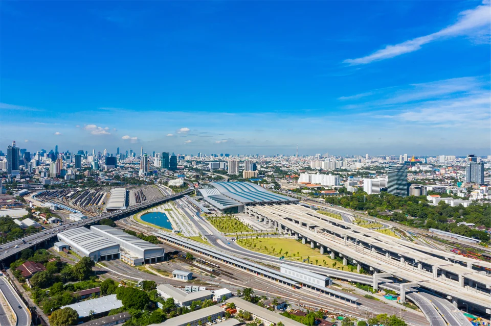 Aerial view of a city transport hub, with elevated rail lines and flyovers converging on a station with a curved striped roof, and a high-rise skyline under a blue sky.