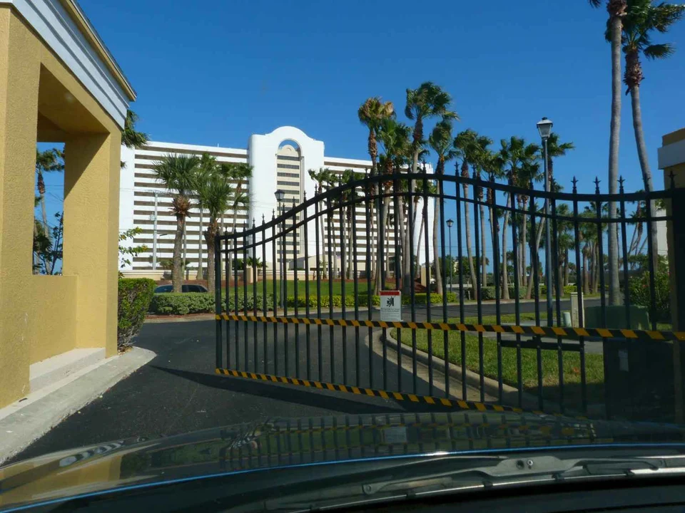 Closed black iron security gate at the entrance to a gated property, seen through a car windscreen, with palm trees and a white high-rise beyond.