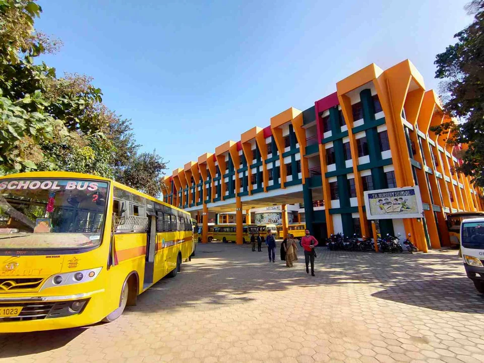 Yellow school bus parked outside a multi-storey school building painted orange, pink and green, with students walking across the paved courtyard.