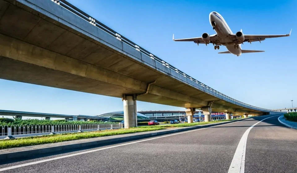 Passenger plane flying low over an elevated flyover, with a low curved-roof building in the distance under a clear blue sky.