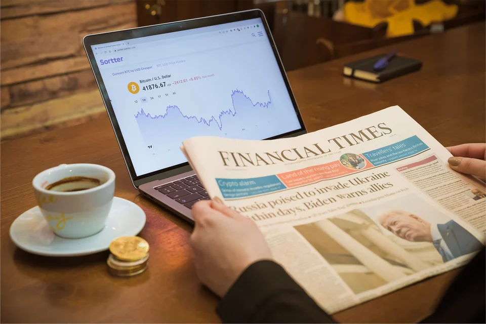 A hand holding a Financial Times newspaper beside a laptop showing a price chart, with a cup of coffee and a small stack of gold coins on a wooden table.