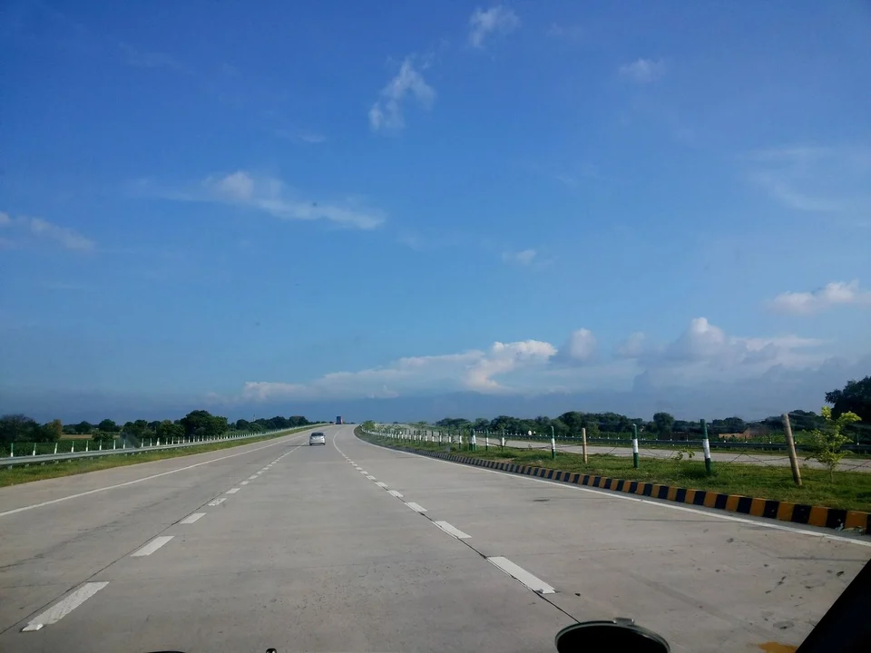 A wide, almost empty expressway stretching to the horizon under a blue sky, with a single car ahead and green verges on both sides.