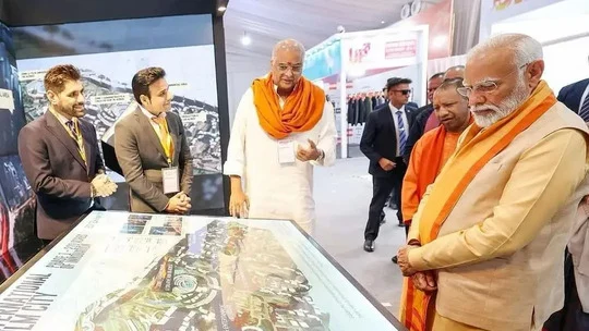 Officials, several wearing saffron scarves, look at an illuminated scale model on a display table while two men in suits explain it.