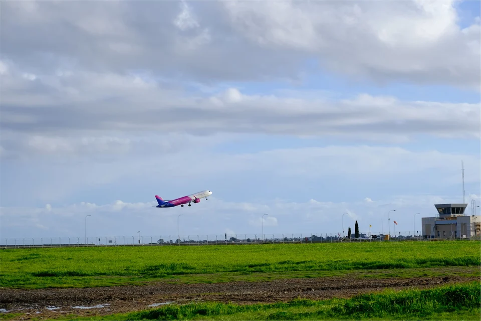 Purple-and-pink passenger jet climbing away from a runway over a green grassy field, with an airport control tower on the right under a cloudy sky.