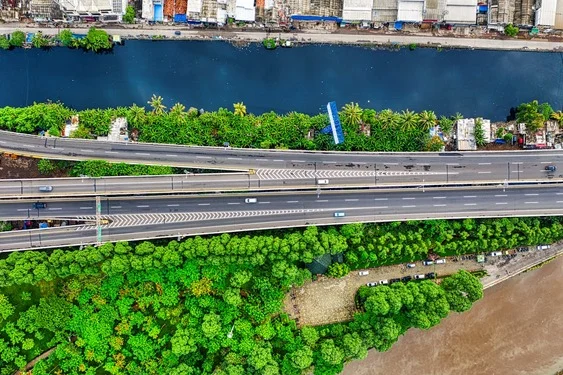 Aerial view of a wide elevated highway carrying light traffic, running between a river and a dense belt of green trees.