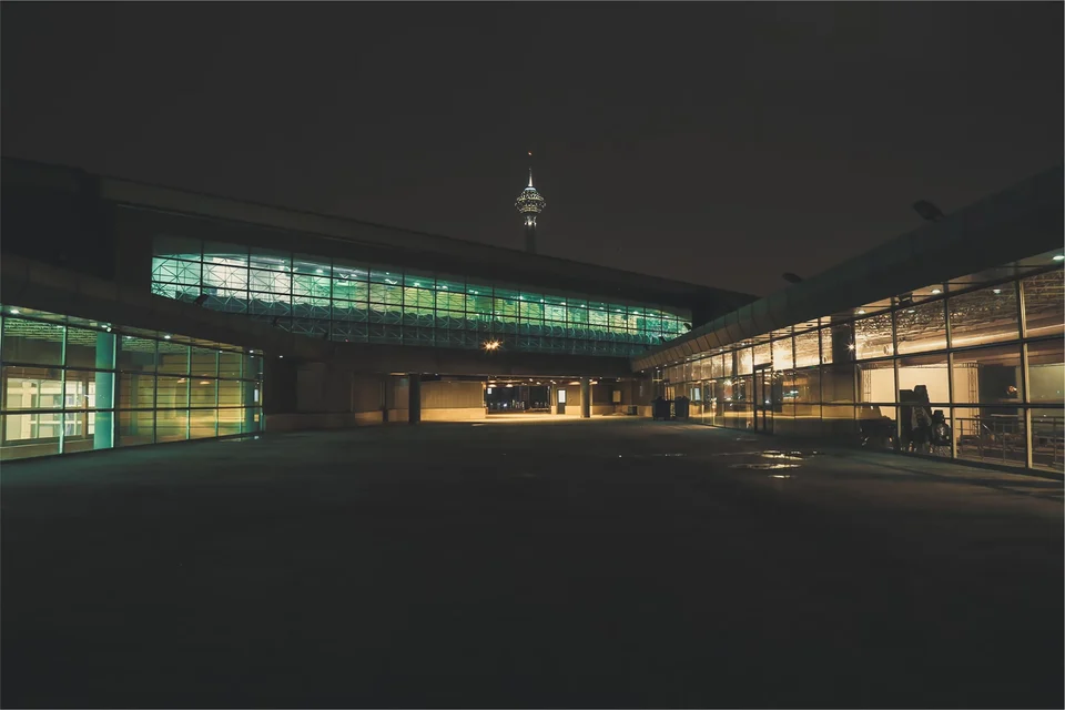 Modern glass-fronted building lit up at night around an empty forecourt, with a lit tower rising behind its roof against a dark sky.