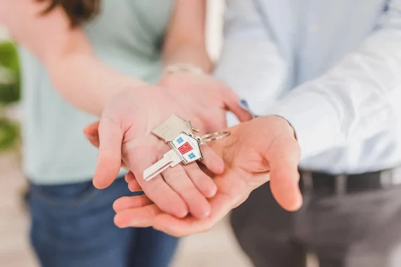 Two people holding out their open hands together, with a house key on a small house-shaped keyring resting in their palms.