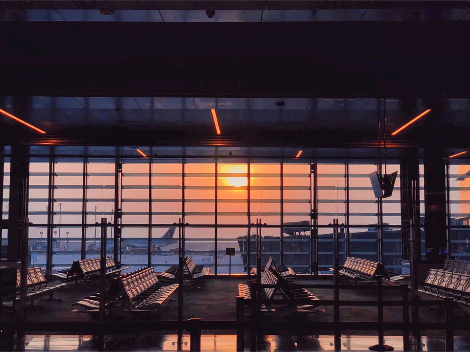Empty airport departure lounge with rows of seats facing tall glass windows, the sun setting over parked aircraft outside.
