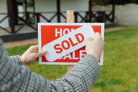 Hands sticking a white 'Sold' label across a red 'Home for Sale' sign on a wooden post in front of a lawn and house.