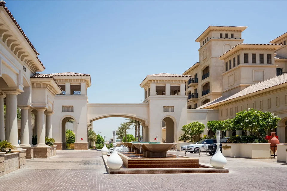 Grand cream-coloured building complex with an arched gateway, columns and tiled roofs around a paved courtyard with a fountain, large white urns and palm trees.