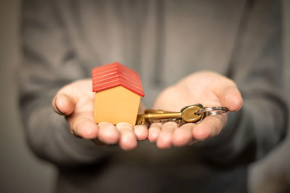 Cupped hands holding out a small toy house with a red roof alongside a bunch of house keys.