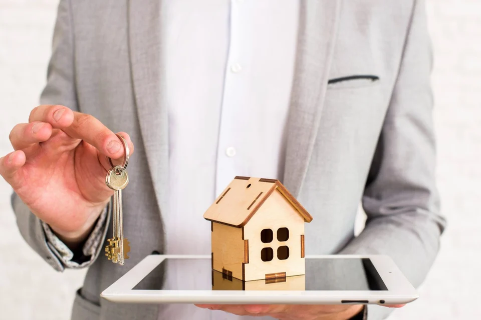 Person in a grey suit holding out a set of keys in one hand and, in the other, a small wooden house model resting on a tablet.
