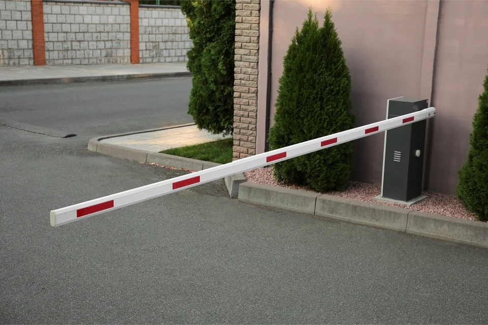 Red-and-white boom barrier lowered across a paved entrance road, with its control box beside trimmed conifer shrubs and a brick wall.
