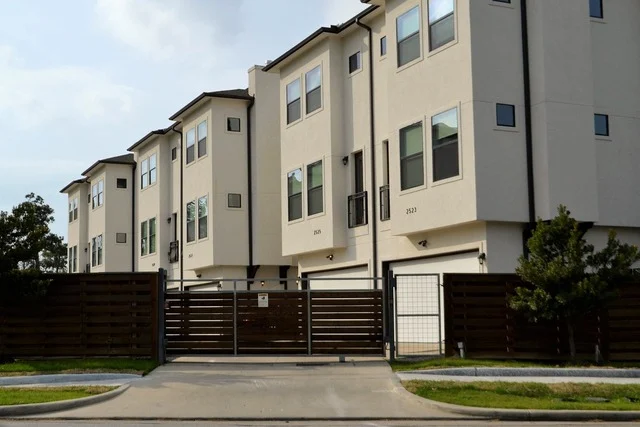 Row of modern three-storey townhouses behind a closed wooden sliding gate and fence, with a paved driveway and lawn in front.