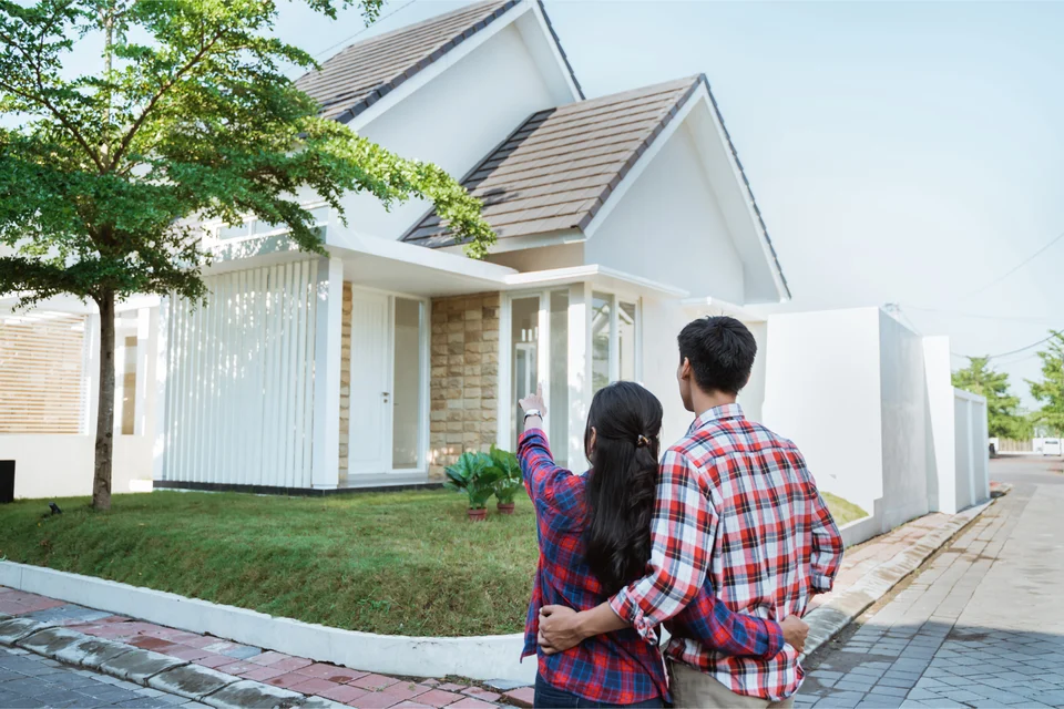 A young couple seen from behind with arms around each other, the woman pointing at a single-storey white house with a grey tiled roof and a front lawn.