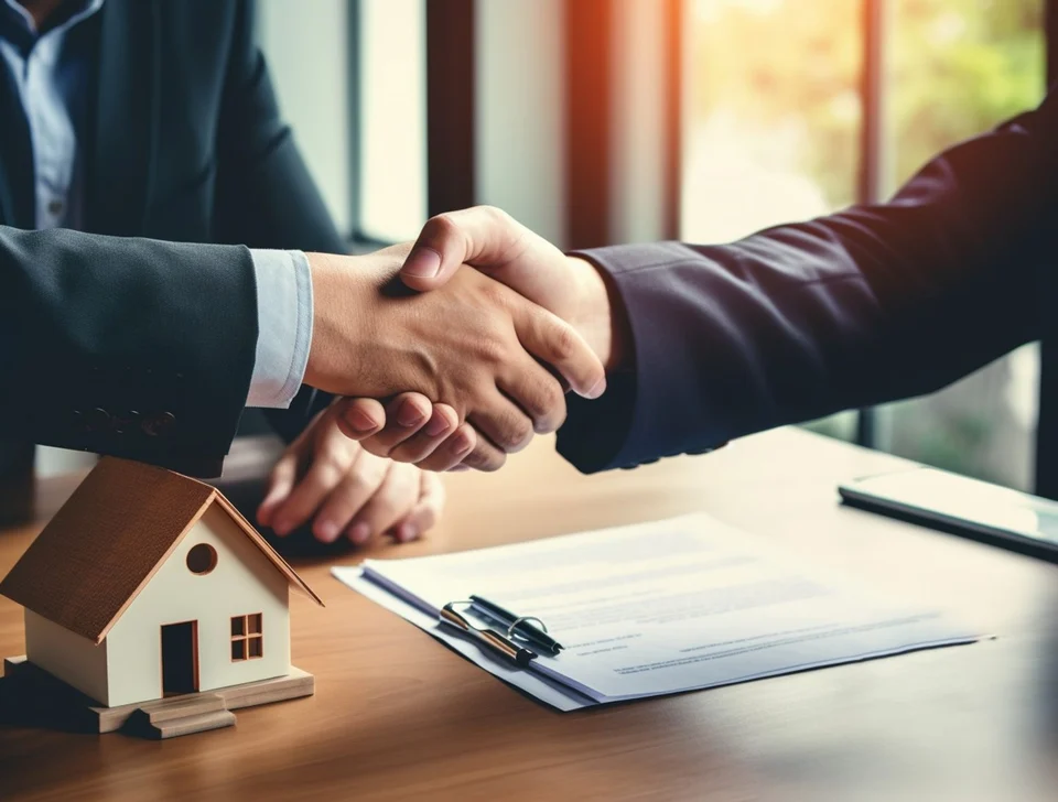 Two people in suits shaking hands over a wooden desk holding a small model house, a document and a pen.