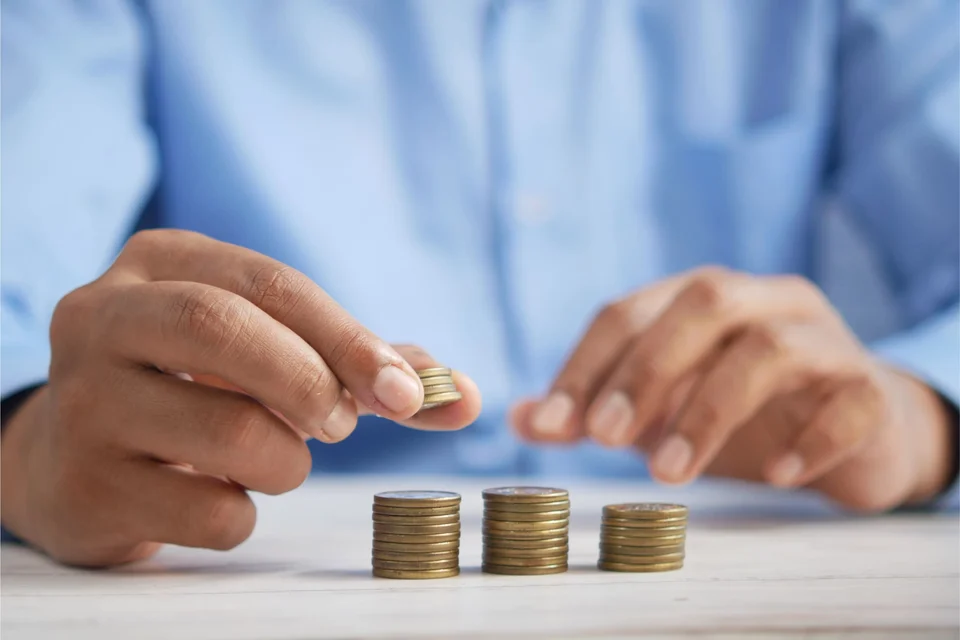 A man in a light blue shirt places a coin on one of three small stacks of coins on a white table.