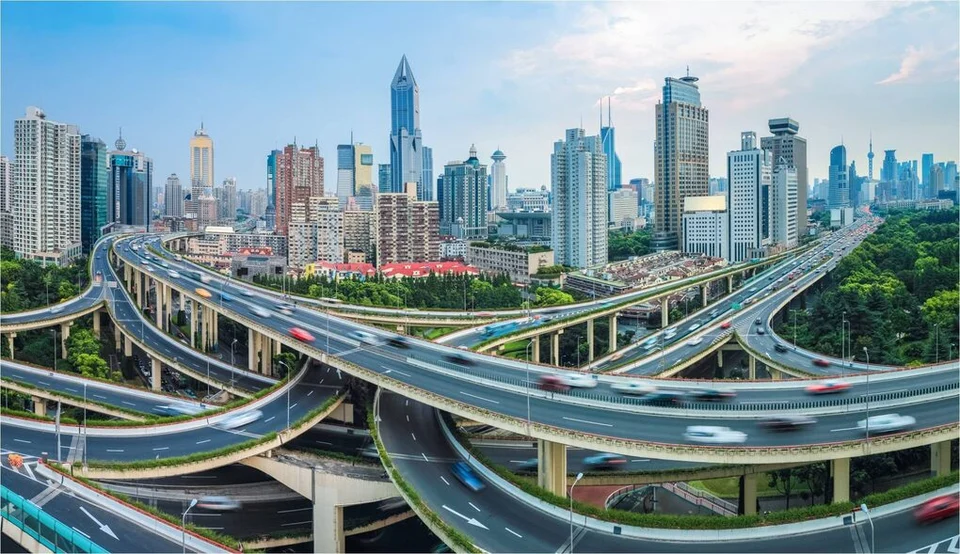 A multi-level highway interchange with curving flyovers full of traffic, set against a dense skyline of high-rise towers.