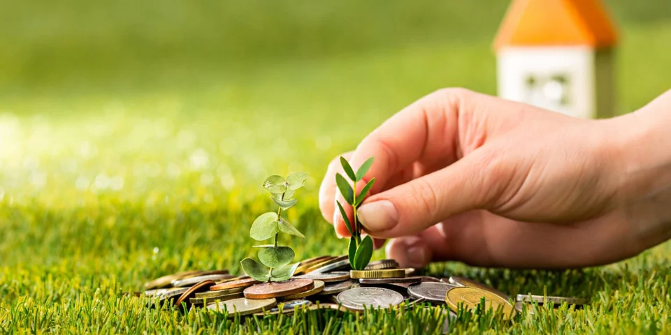 A hand planting a small seedling among scattered coins on green grass, with a small model house blurred in the background.