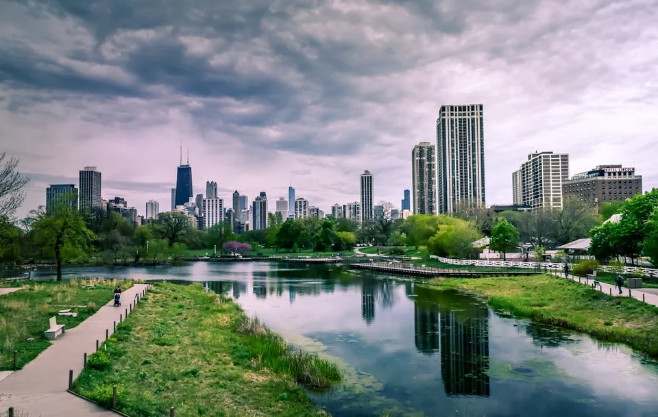 City skyline of high-rise towers reflected in a park pond, with grassy banks, a footpath and trees under a cloudy sky.