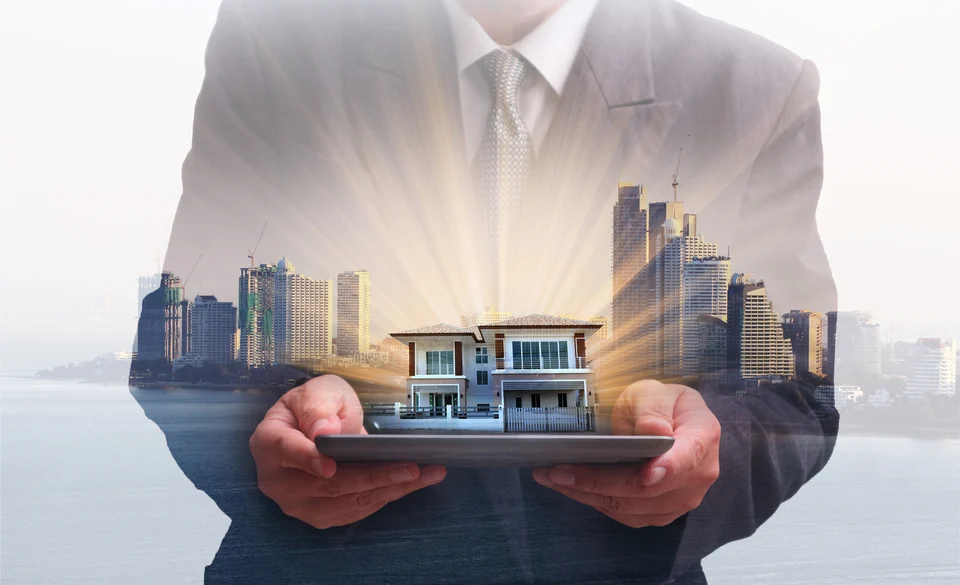 Double-exposure image of a man in a suit holding a tablet with a glowing model house on it, overlaid with a skyline of high-rise towers by the water.