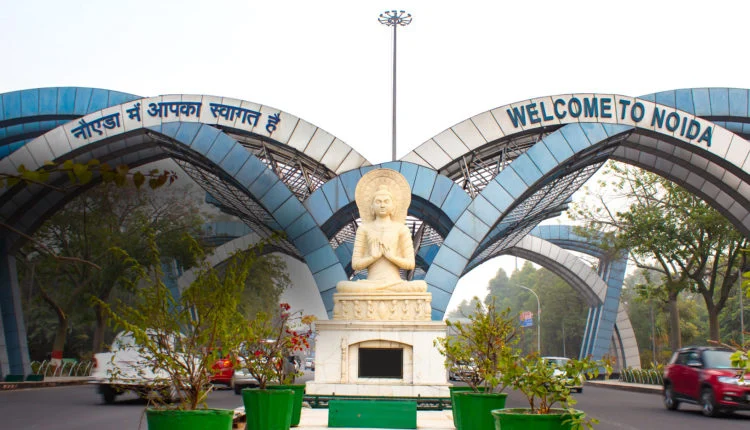 Noida entrance gate with blue arches reading 'Welcome to Noida' in English and Hindi, a seated Buddha statue in the centre and potted plants in front.