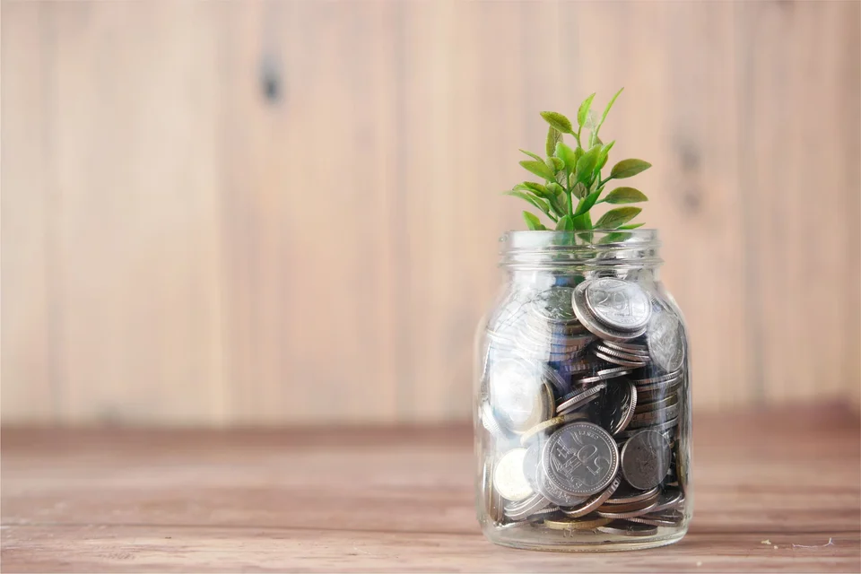 A small green plant growing out of a glass jar filled with coins, on a wooden table.