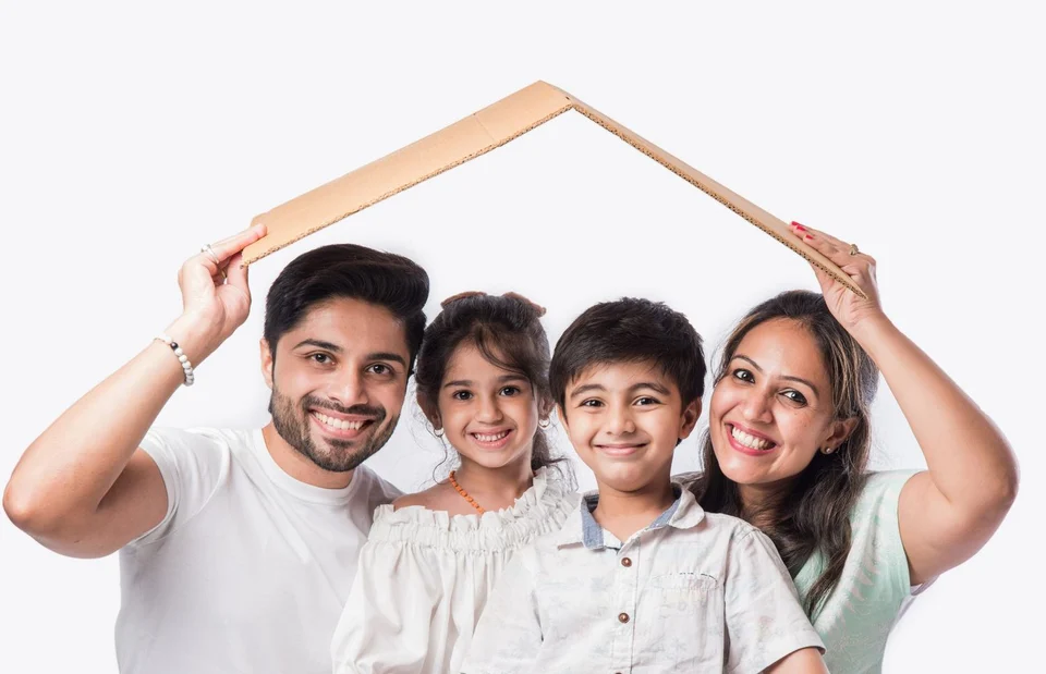 A smiling family of four, two parents with a young daughter and son, with the parents holding a cardboard roof shape above their heads.