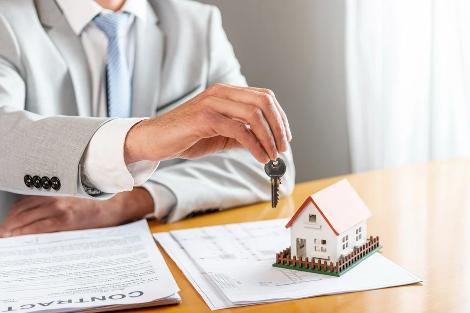 A man in a grey suit holds up a house key over a desk with a small model house, a floor plan and a contract.