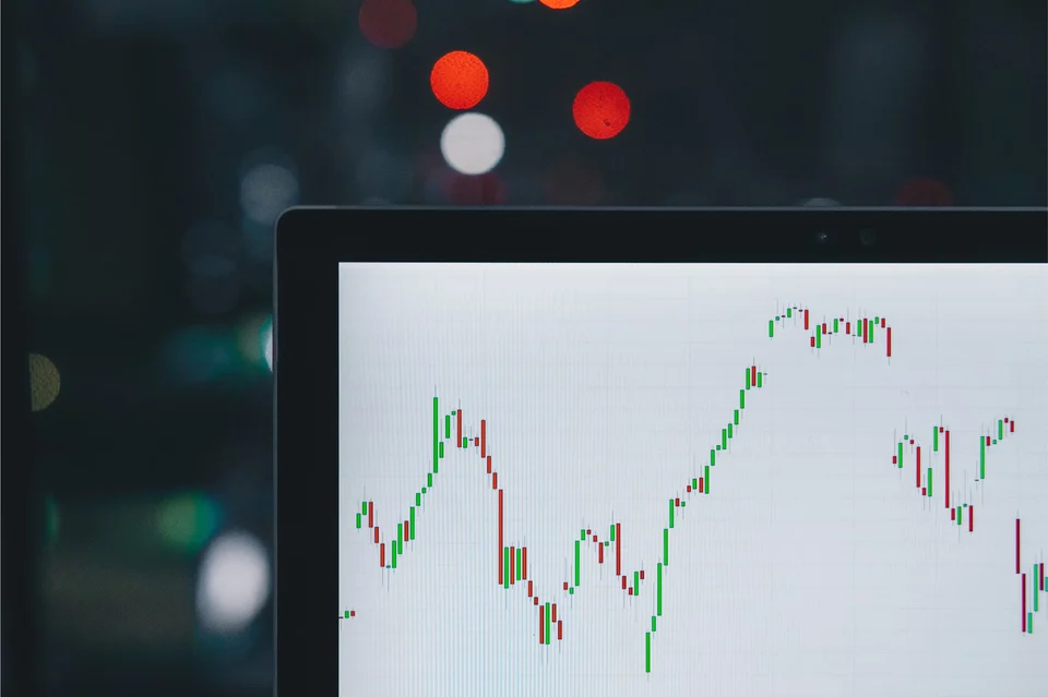 Laptop screen showing a red and green candlestick price chart, with blurred city lights in the dark background.