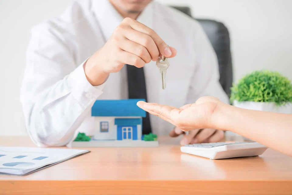 Man in a white shirt and tie at a desk handing a house key to an outstretched hand, beside a small blue house model and a calculator.
