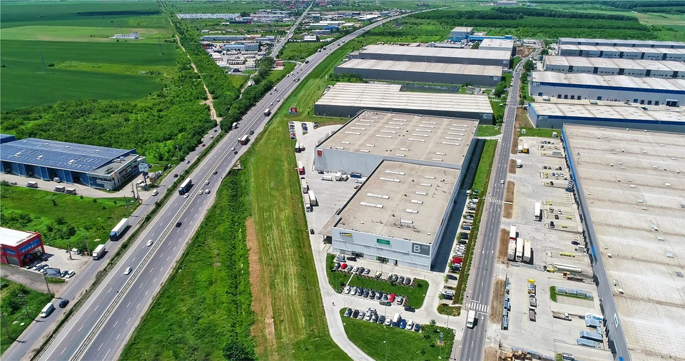 Aerial view of a large logistics park with rows of warehouse buildings, truck yards and parking beside a multi-lane highway, surrounded by green fields.