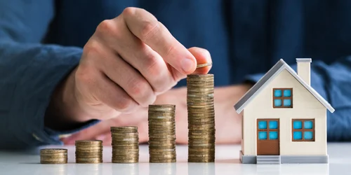 A hand placing a coin on the tallest of several rising coin stacks, next to a small model house with blue windows.