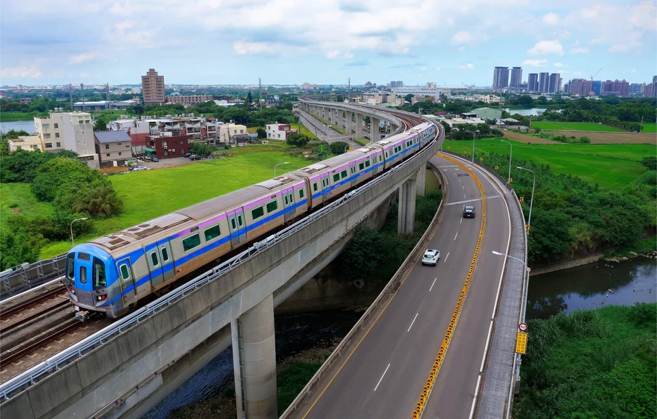 An elevated metro train on a curving viaduct beside an elevated road with cars, crossing green fields with a town and high-rise towers in the distance.