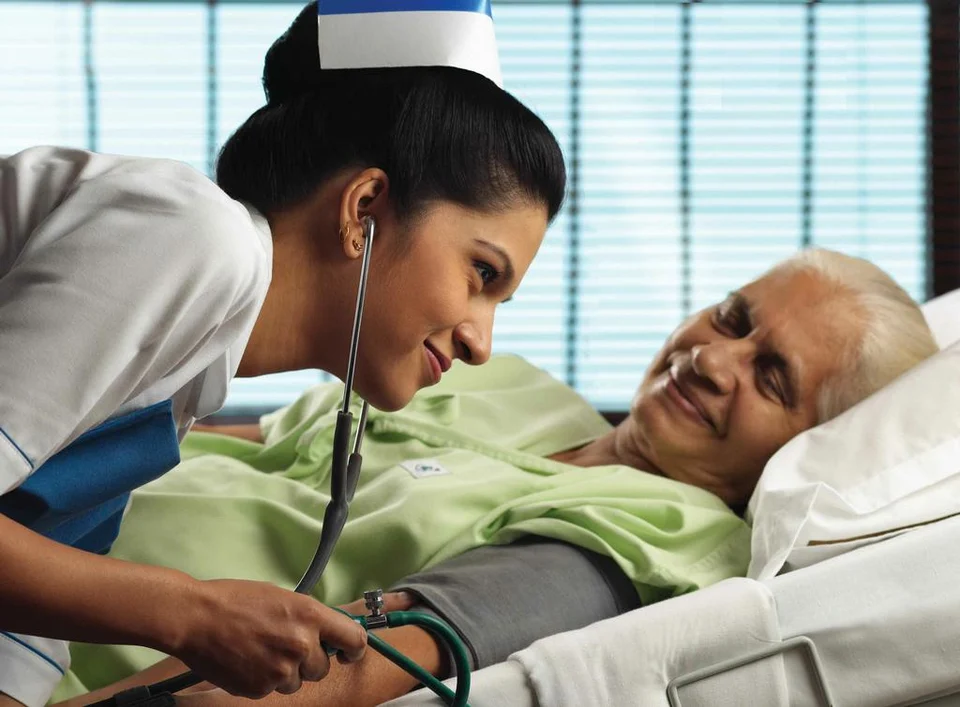 A nurse in a white cap checks the blood pressure of a smiling elderly patient lying in a hospital bed.
