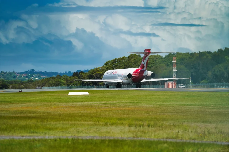 White passenger jet with a red tail on a runway, with a wide strip of green grass in front and trees and dark storm clouds behind.