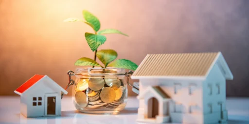 Glass jar filled with coins with a green seedling growing out of it, placed between two small model houses.