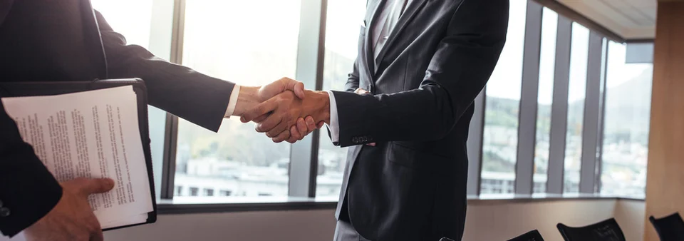 Two people in business suits shaking hands in an office with tall windows, one holding a clipboard of printed documents.