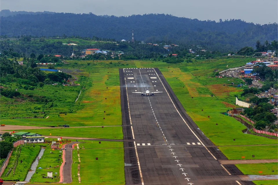 A passenger jet on a long airport runway cut through green fields, with houses on the slopes and forested hills behind.